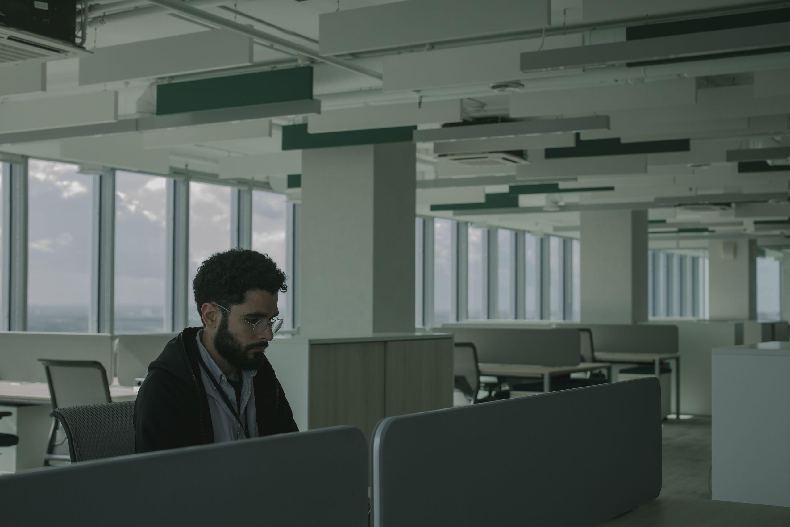 Man with glasses working alone at a desk in a spacious open-plan office with large windows