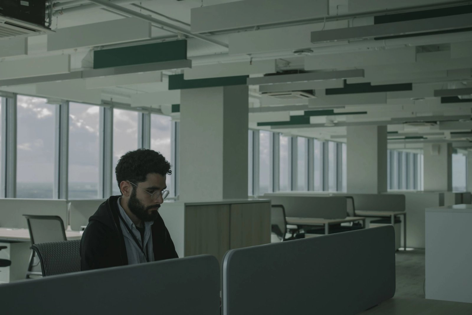 Mann mit Brille arbeitet allein an einem Schreibtisch in einem weitläufigen Großraumbüro mit Fensterfront