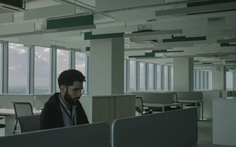 Man with glasses working alone at a desk in a spacious open-plan office with large windows