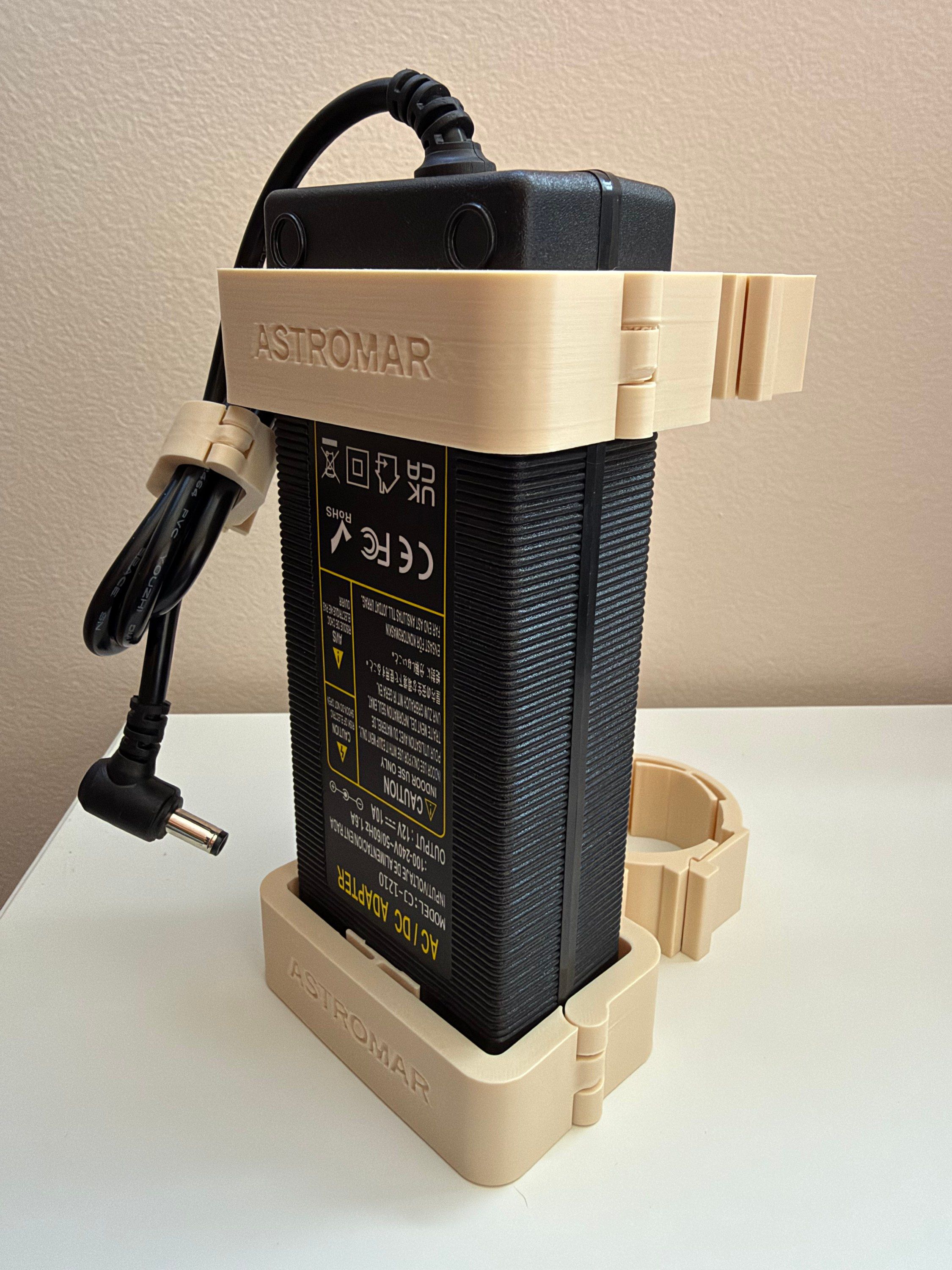 The same holder from the side, gripping a black 12V 10A AC/DC power brick, with the bottom clamp, a hinged cable clip and ASTROMAR embossed on the plastic
