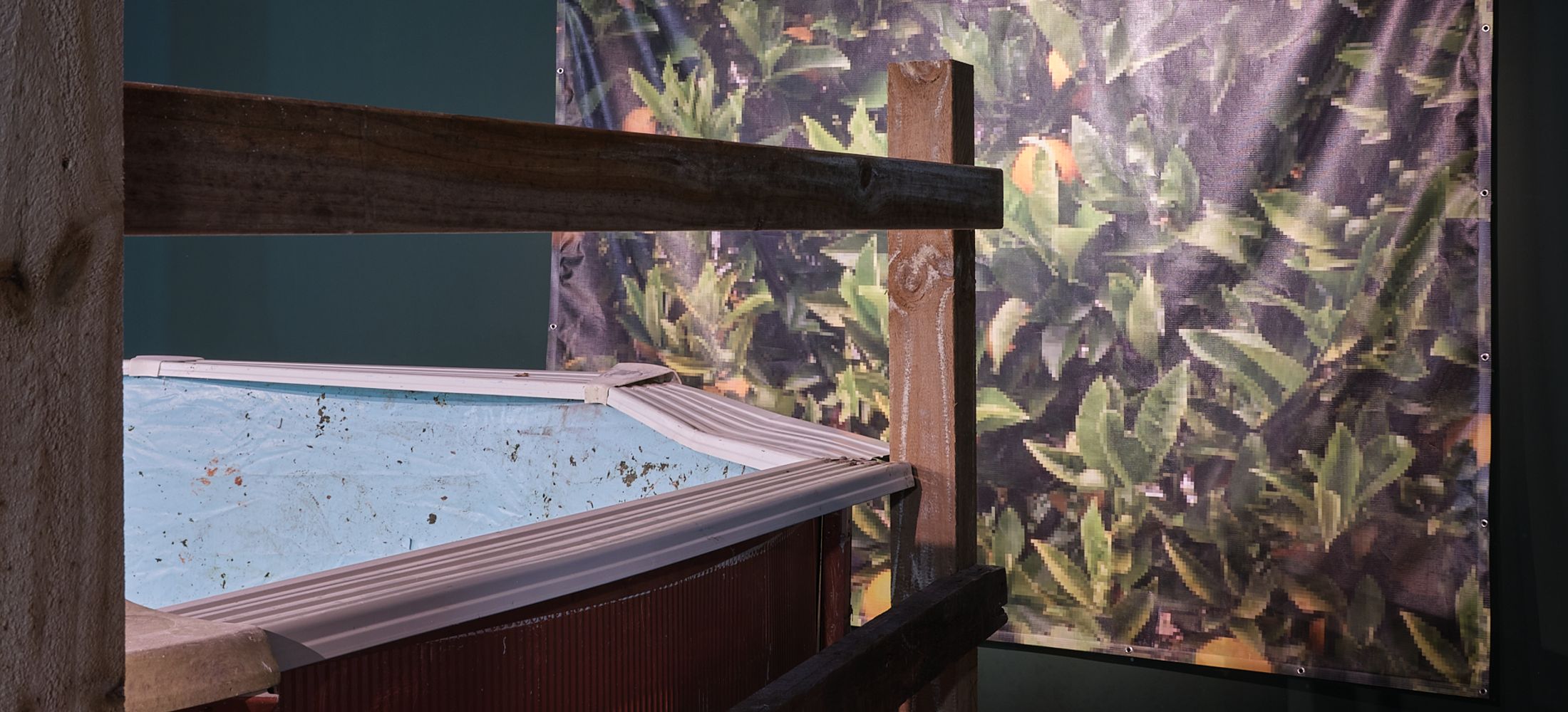 An aspect of the Vacant Possession Exhibition featuring an above ground pool with incomplete framing and fallen leaves. A digital print of citrus trees obscures the dark wall behind. 