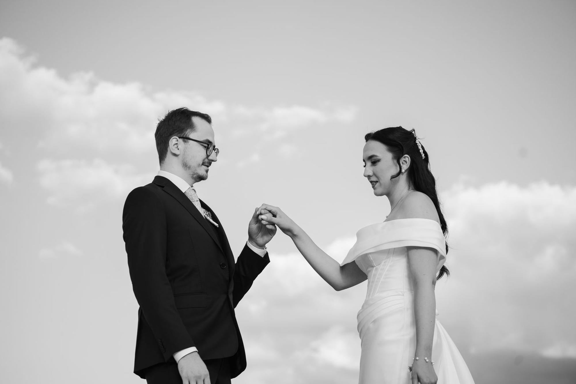 Black and white photo of the groom holding the bride’s hand against a cloudy sky