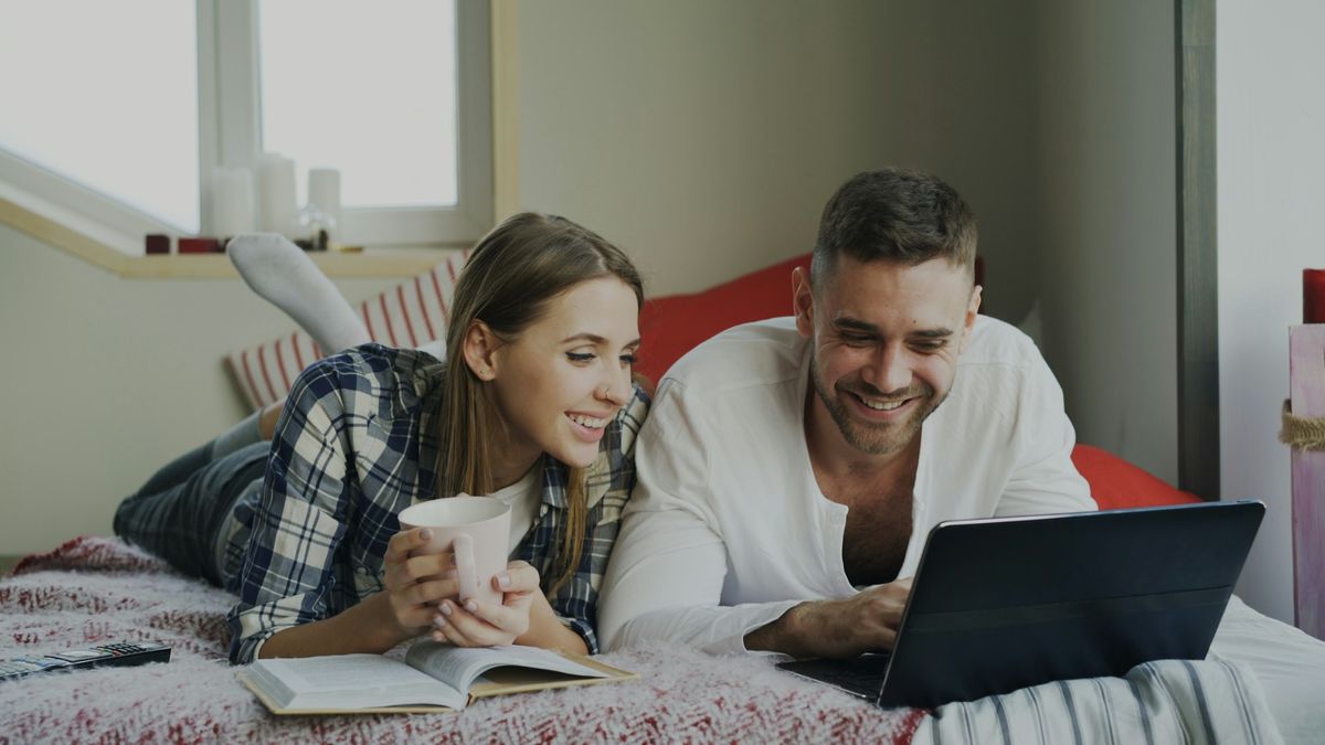 Smiling man and woman talking and lying in bed reading book and using laptop computer in the morning