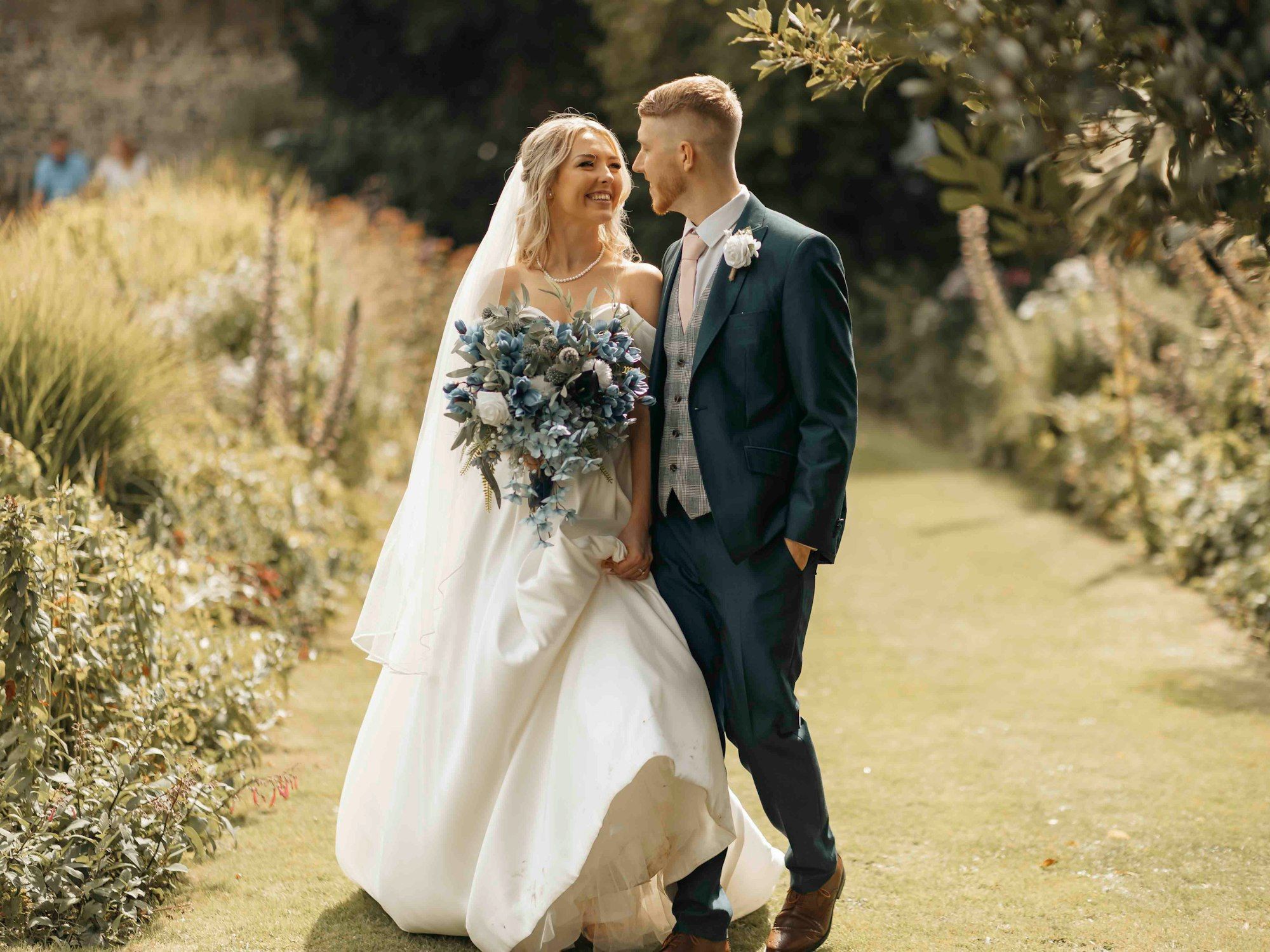 Bride and groom walking together in a garden, bride holding a blue-toned bouquet