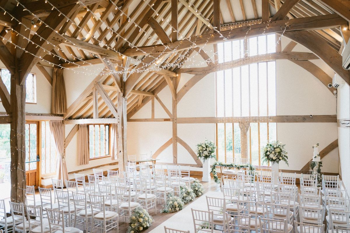 Timber-framed barn ceremony room at Harper Weddings with fairy lights and white chiavari chairs