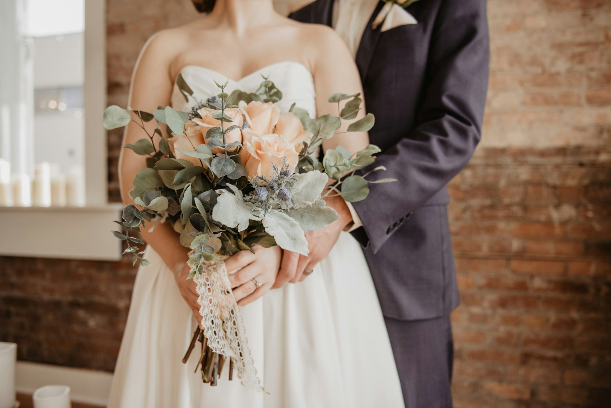 Bride and groom close up holding a peach and eucalyptus bouquet against a brick wall