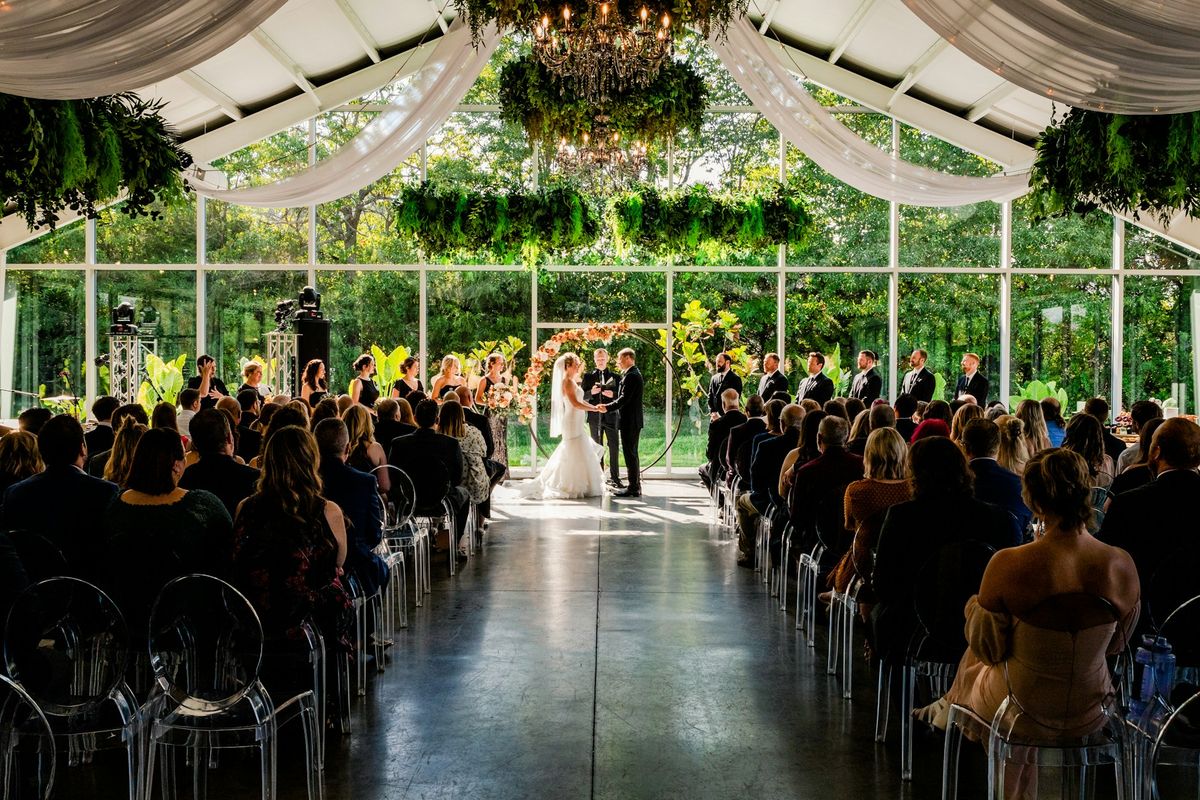 Wedding ceremony inside a glass pavilion decorated with hanging greenery