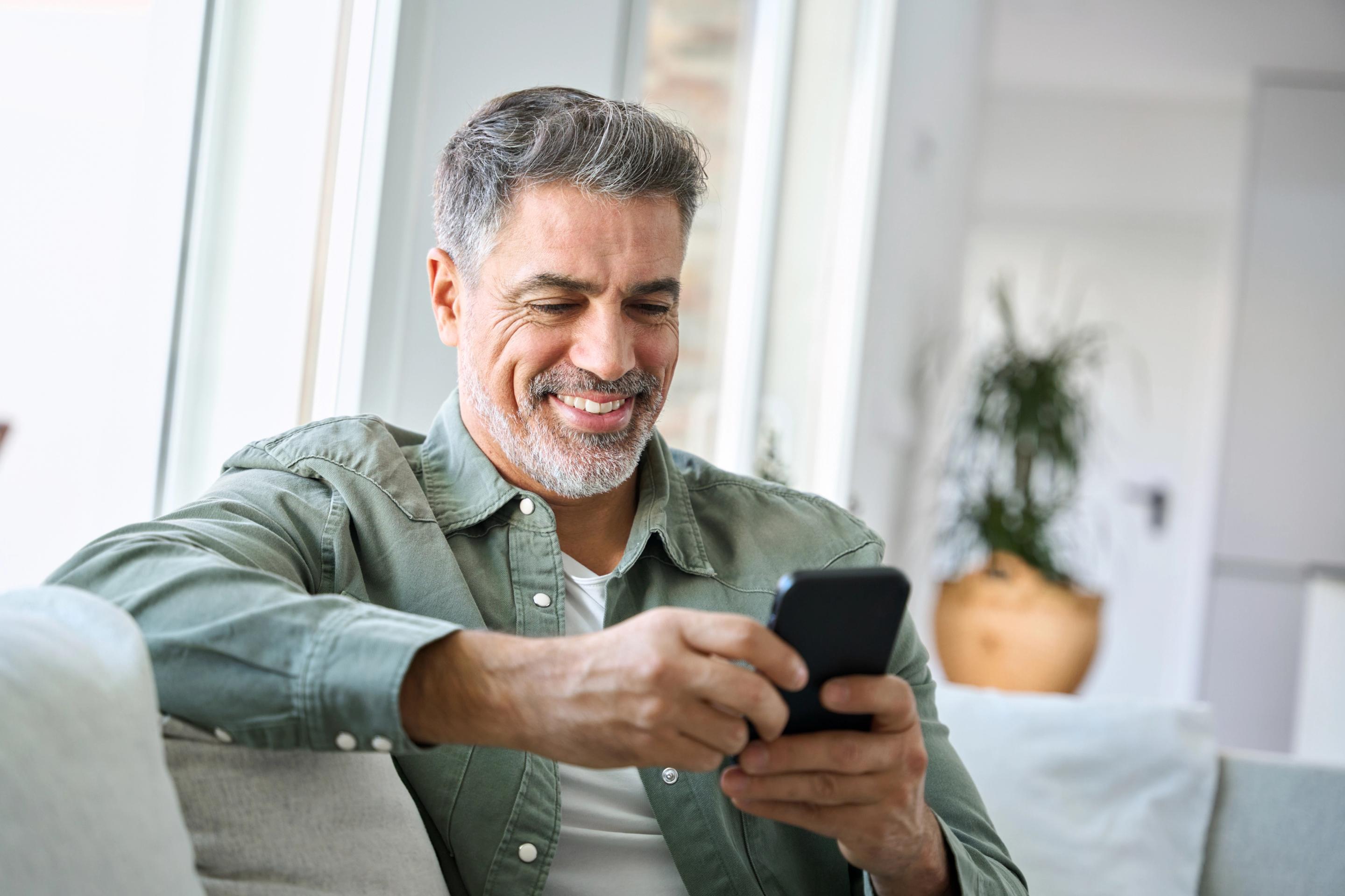 Man in green shirt sitting down on couch smiling at phone in hands.