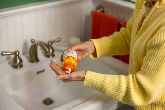 Woman in yellow sweater dispensing two pills into hand from pill bottle