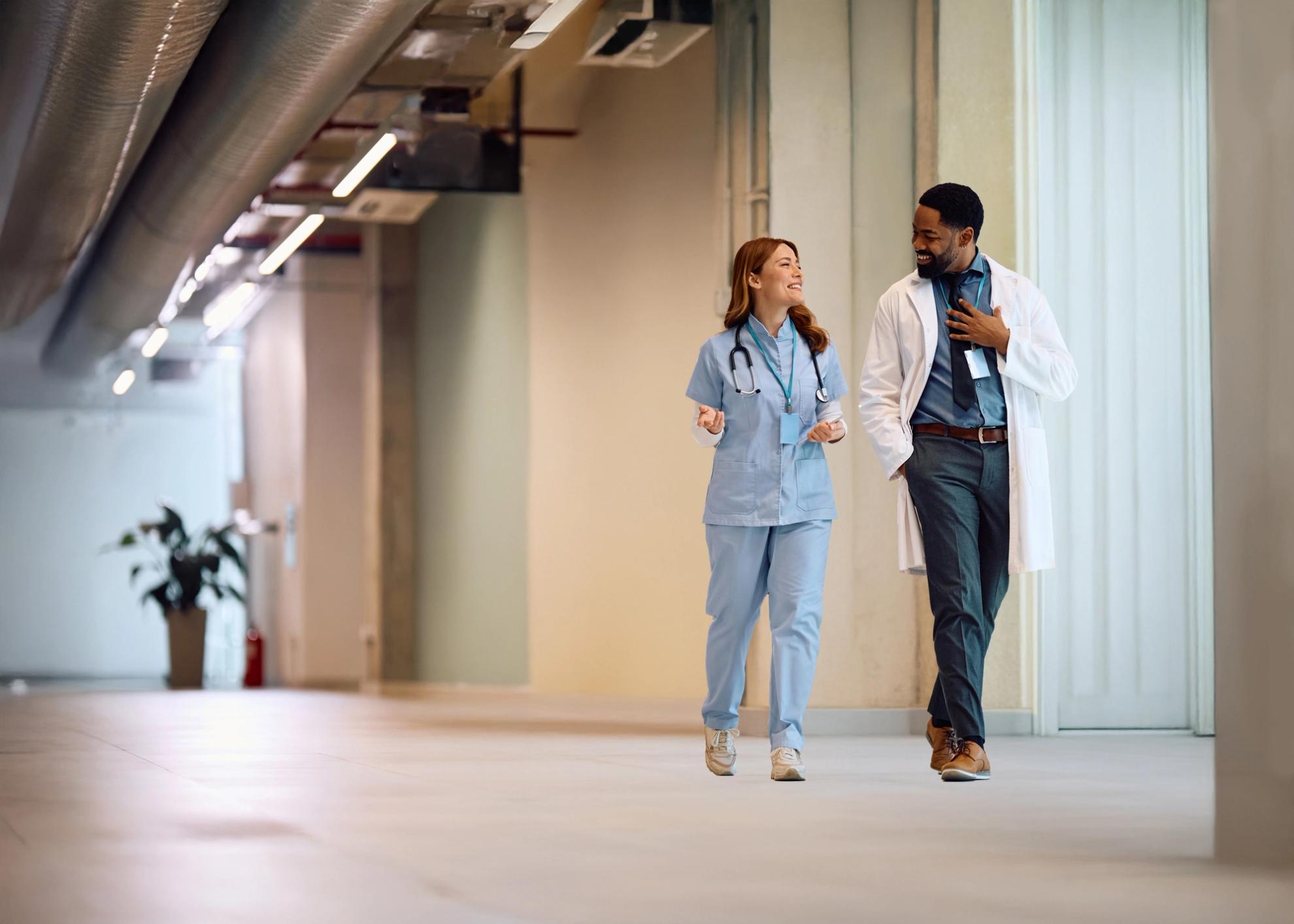 Two healthcare professionals, one in scrubs and one in a lab coat, walk and talk in a hospital