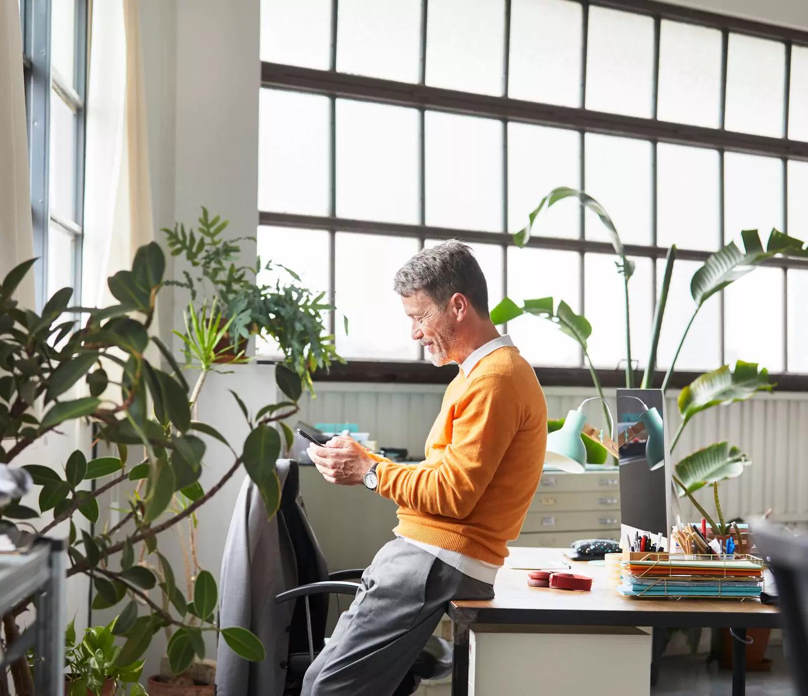 Man in orange sweater leaned against desk looking at phone