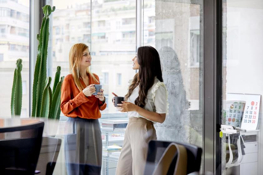 Two women coworkers in office drinking coffee out of mugs and talking