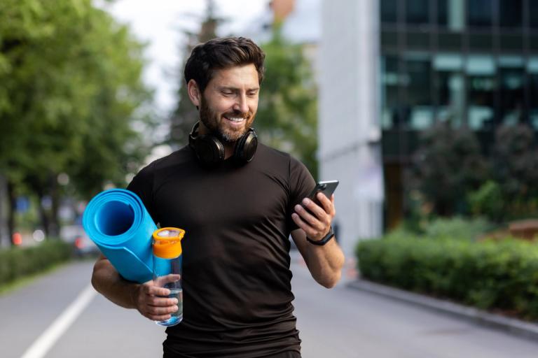Man in his 30s on his way to the gym with a yoga mat, water bottle, and headphones around his neck, looking down at his phone.