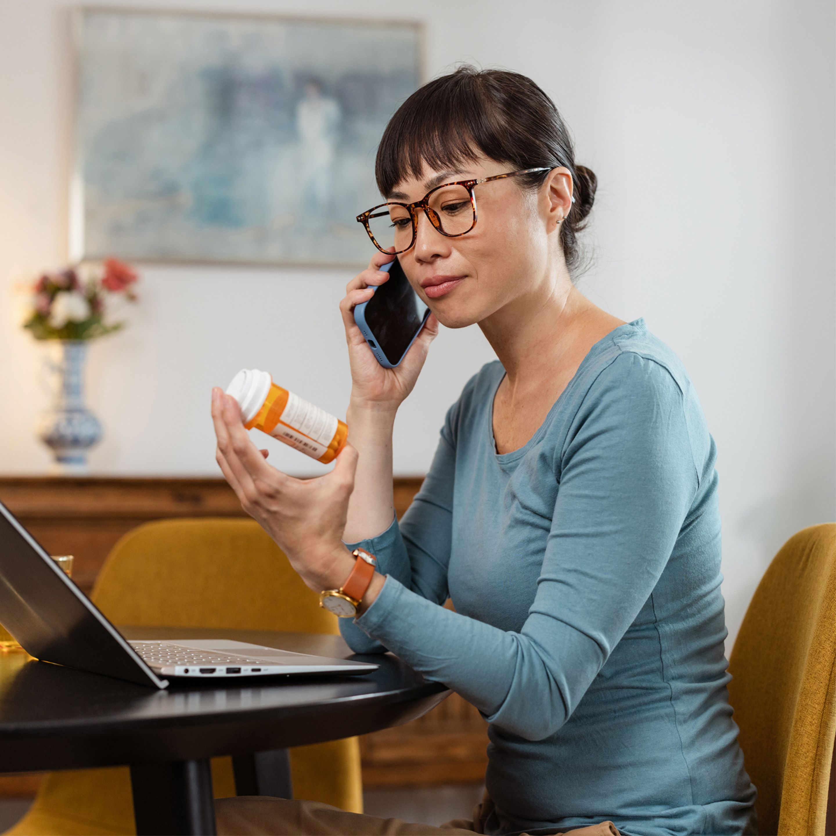 Woman in blue shirt with glasses looking at medicine bottle while on the phone