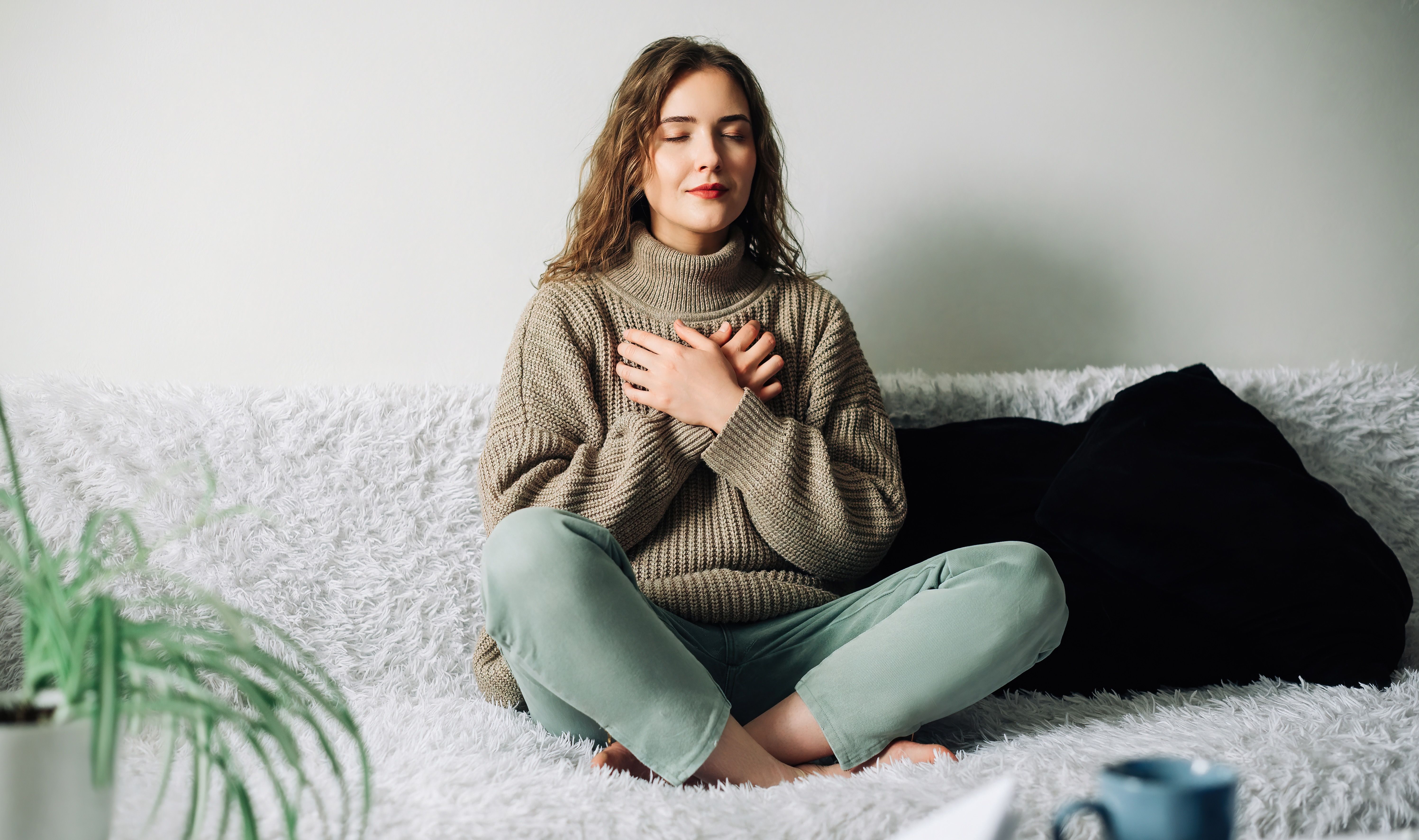 Women in comfy clothes in her home doing breathing exercises. She's sitting with her legs criss-crossed and her arms crossed over her chest. Her eyes are closed taking deep breaths.