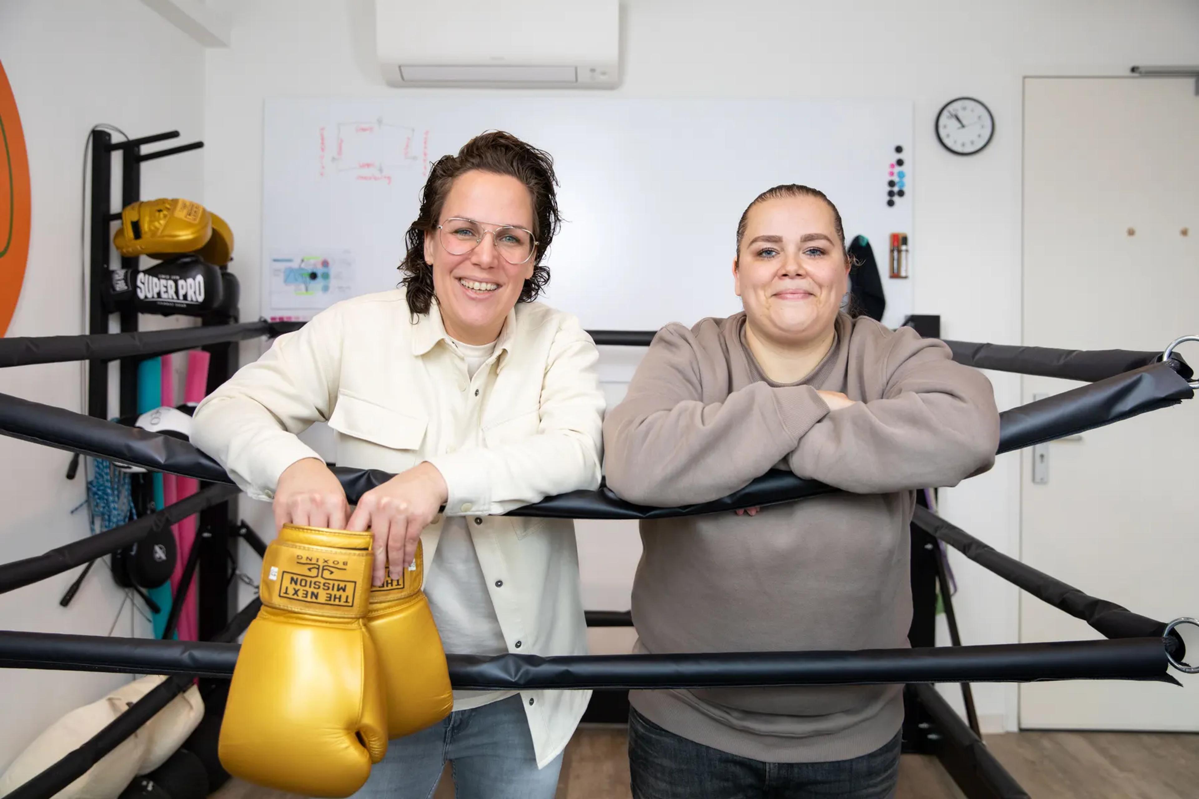 Samantha en een cliënt leunen lachend op de touwen van de boksring, gouden handschoenen ervoor