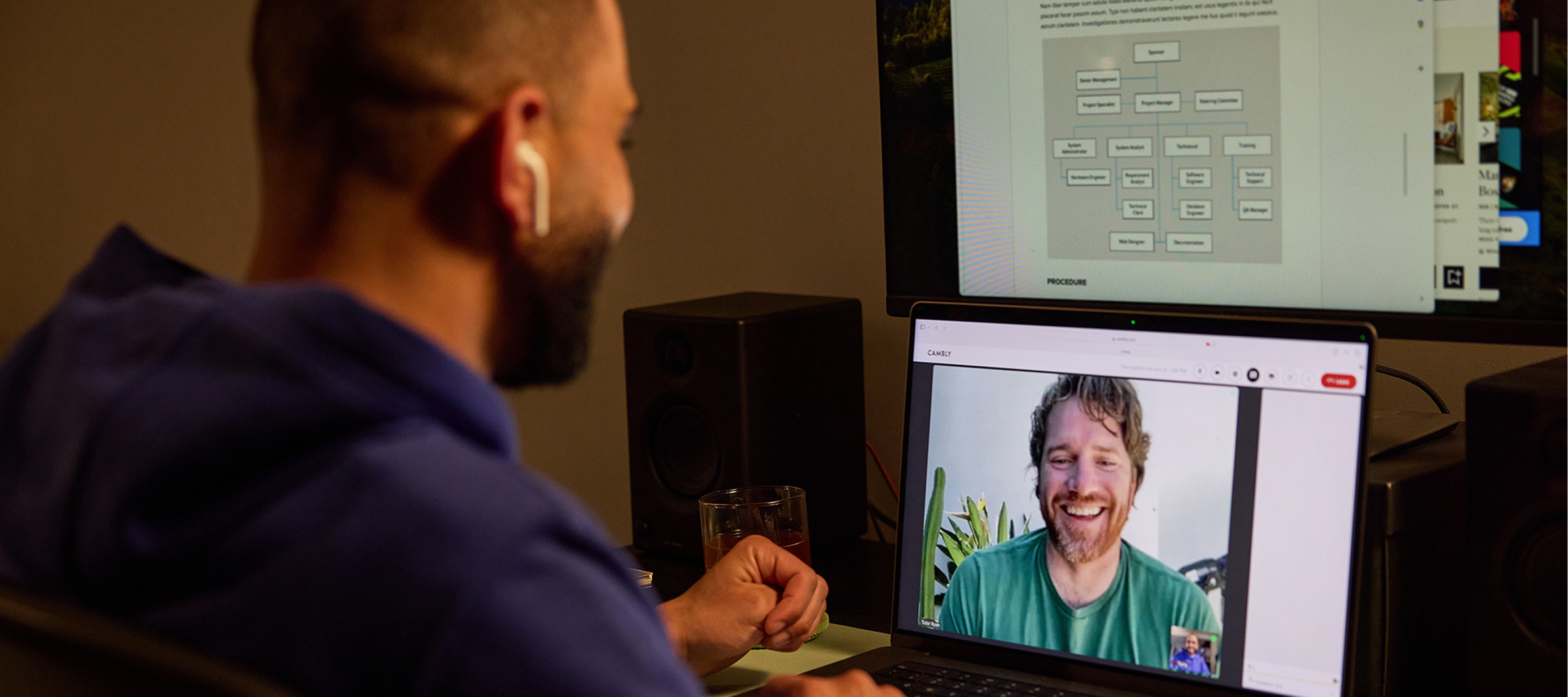 Profissional em uma aula de inglês para negócios no Cambly pelo notebook, com o tutor nativo sorrindo na tela e um organograma da empresa aberto no monitor acima.