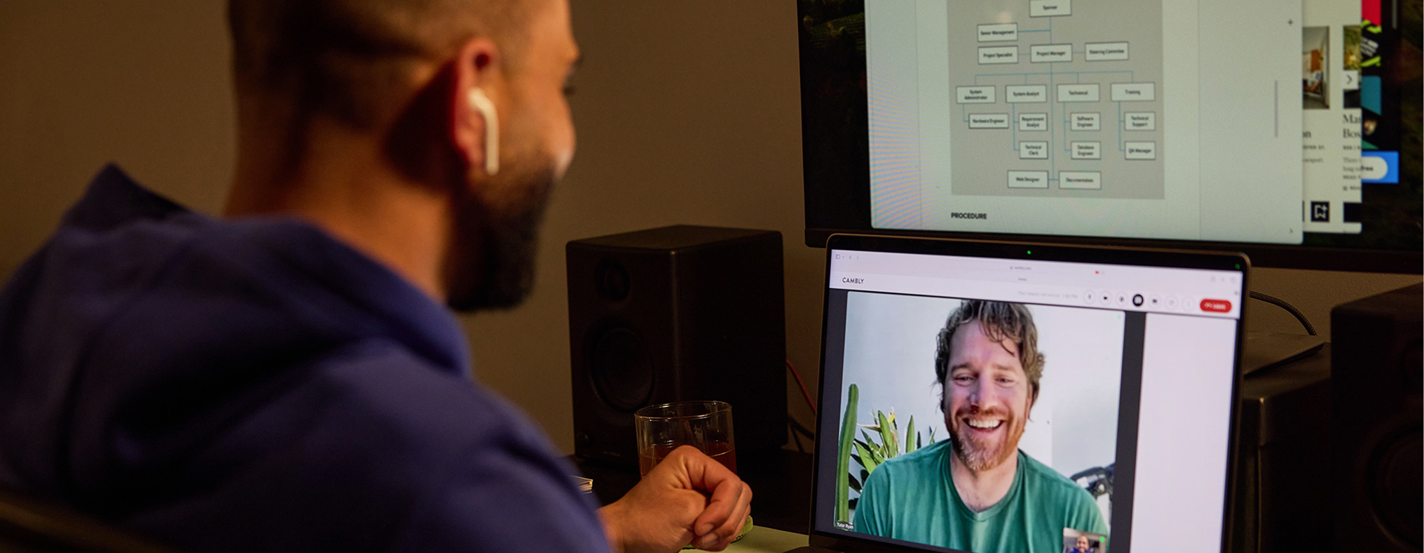 Profissional em uma aula de inglês para negócios no Cambly pelo notebook, com o tutor nativo sorrindo na tela e um organograma da empresa aberto no monitor acima.
