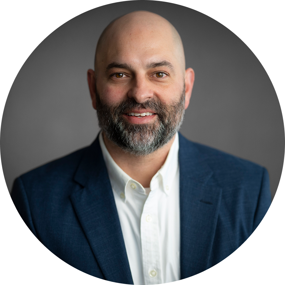 Headshot of a bald man with a beard smiling, wearing a blue blazer and white shirt against a gray background.
