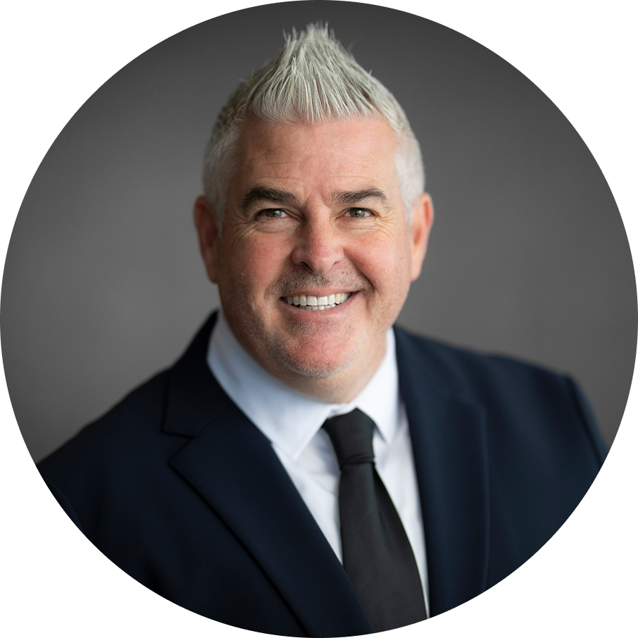 Man in suit and tie with silver hair, smiling. Dark gray background. Head and shoulders shot.