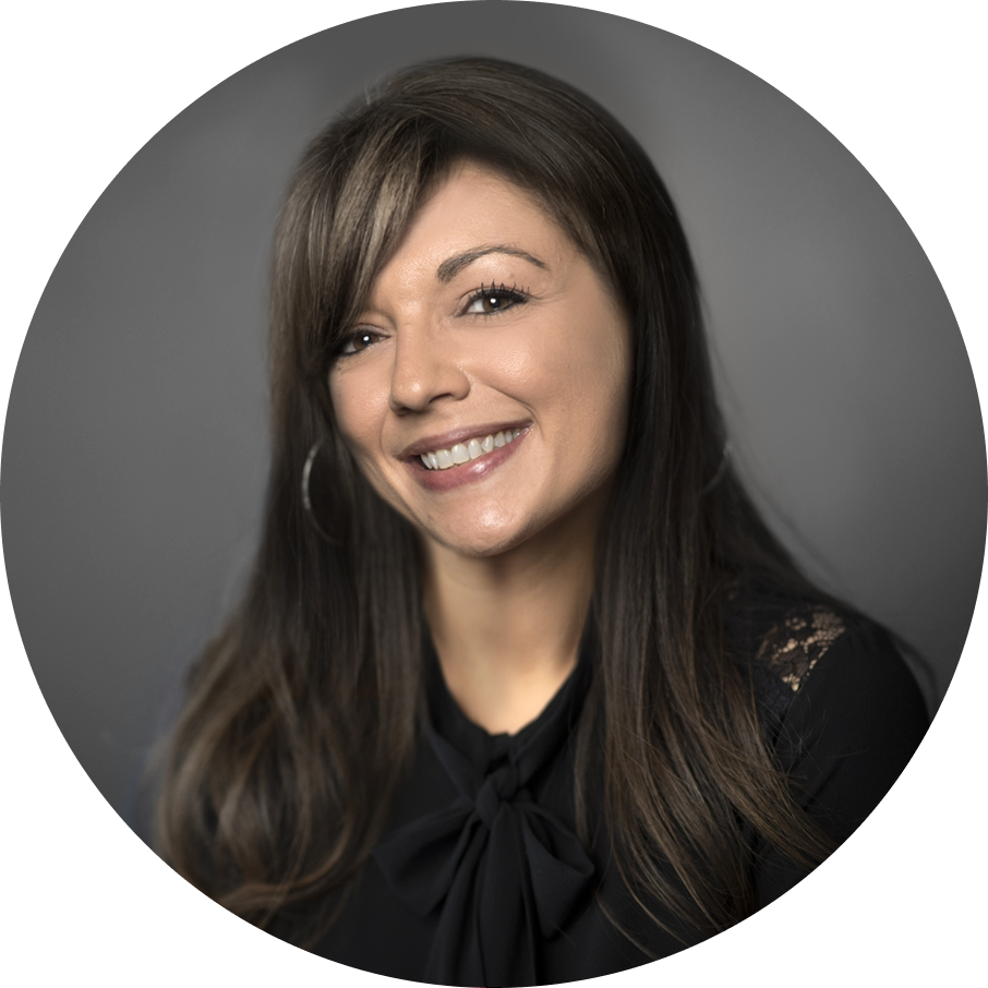 Smiling woman with long brown hair, bangs, hoop earrings, black top with a bow, and lace details. Gray background.