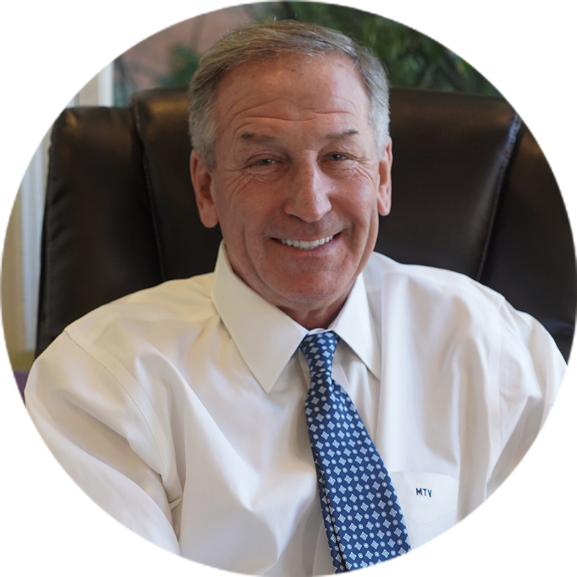 Smiling older man in a white shirt and blue tie sits in a leather office chair.