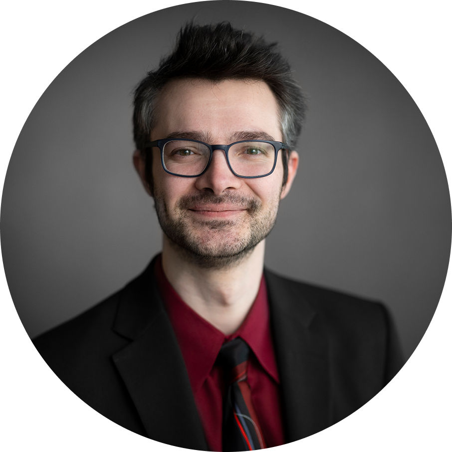 Smiling man with glasses, in black suit and red shirt, against a gray background.