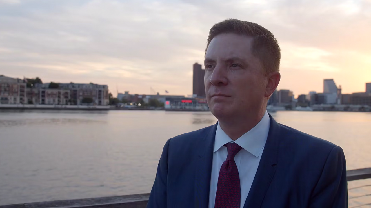 Man in suit with harbor and city skyline at sunset in background.