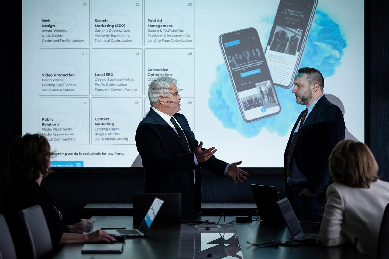 Business meeting in a conference room with two men presenting to colleagues. A website design presentation is displayed.