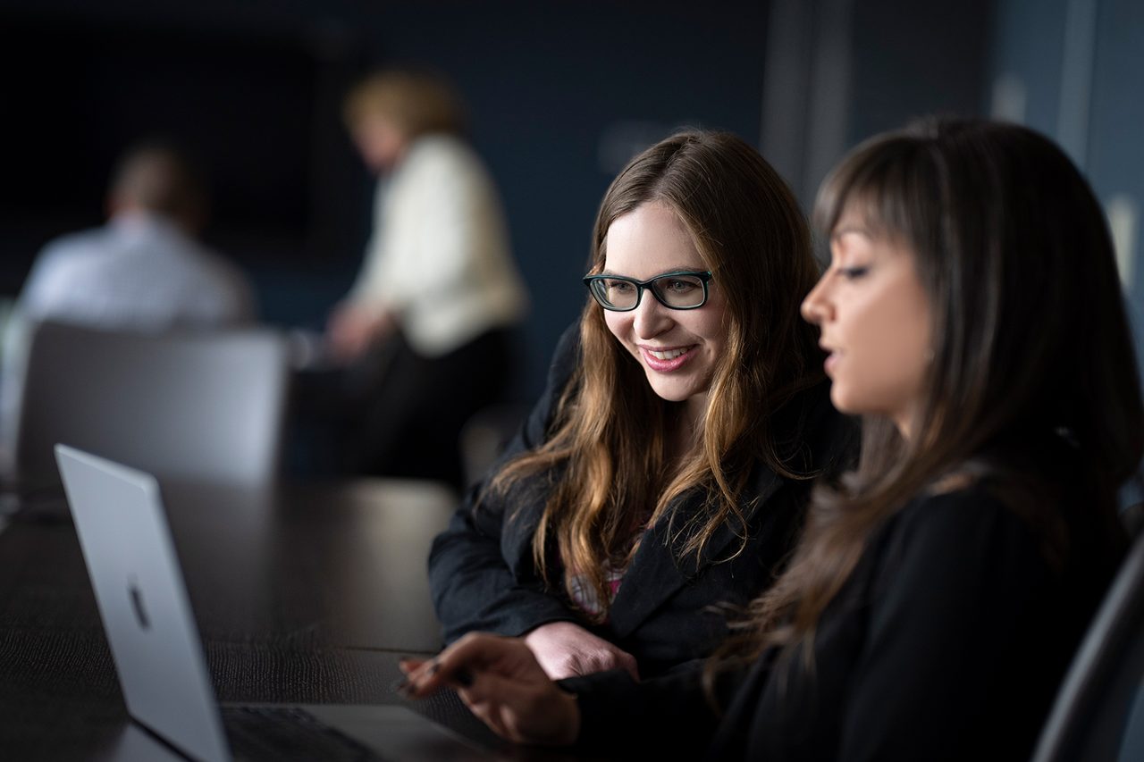 Two businesswomen at a table looking at a laptop, discussing its contents, in a modern office setting.