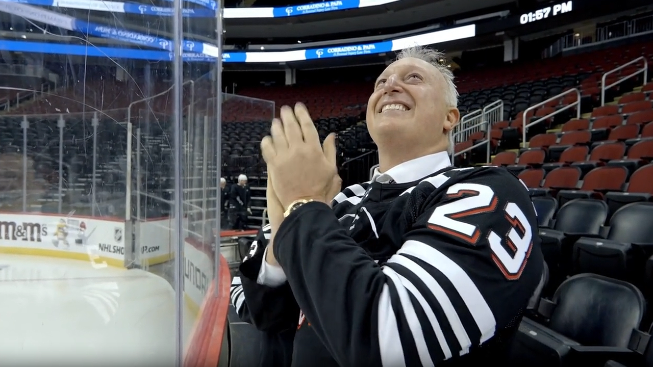 Man in a hockey jersey claps at rinkside. He’s smiling, with empty stadium seats behind him. Game time is 1:57 PM.