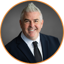Headshot of a smiling man with gray, styled hair, wearing a suit and tie, set against a gray background within an orange c...