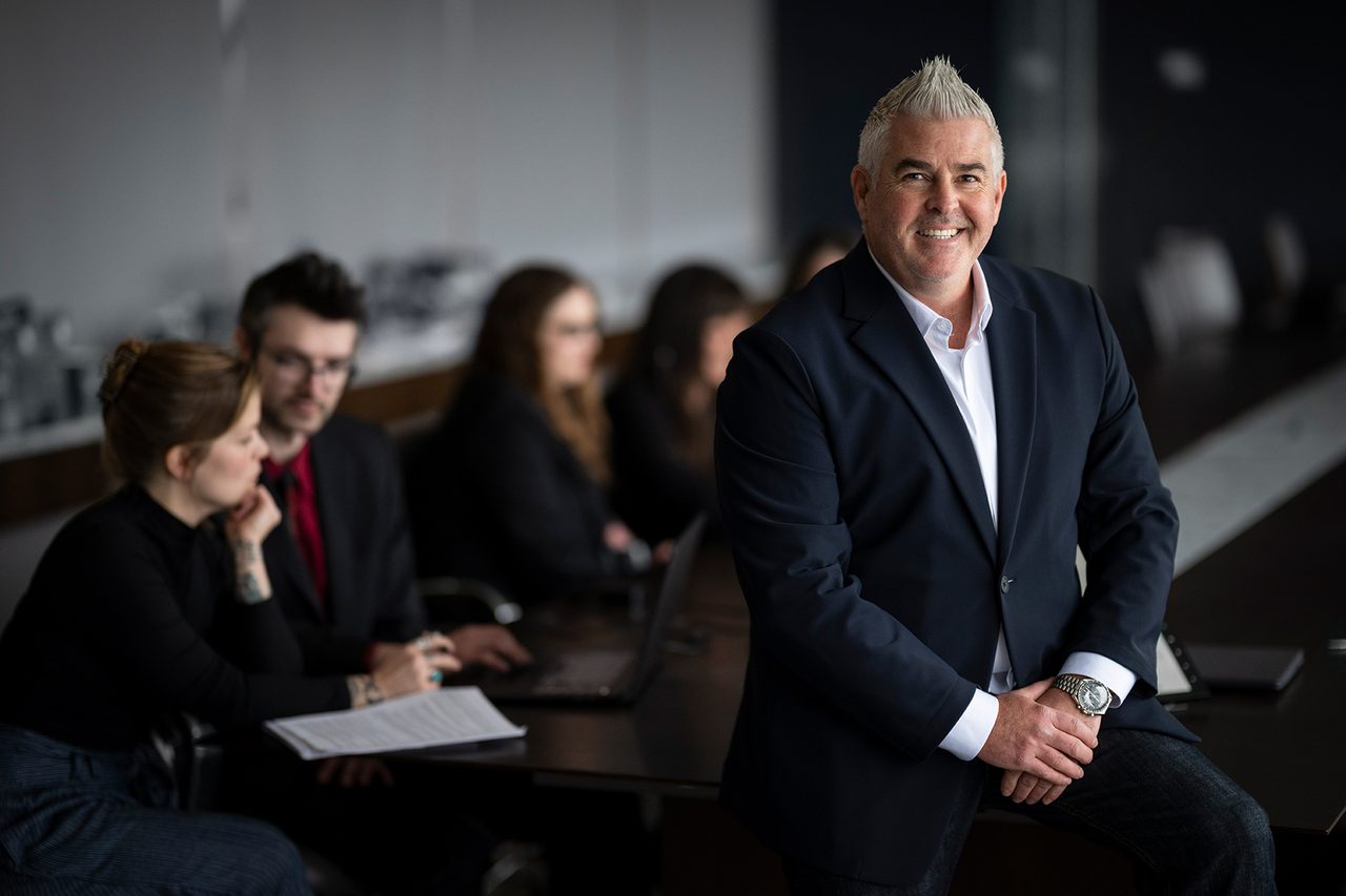 Man with spiky hair in suit smiles, leaning on conference table with blurry people in the background.