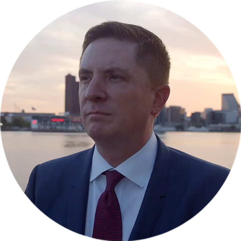 Man in suit & tie stands before waterfront city at sunset.