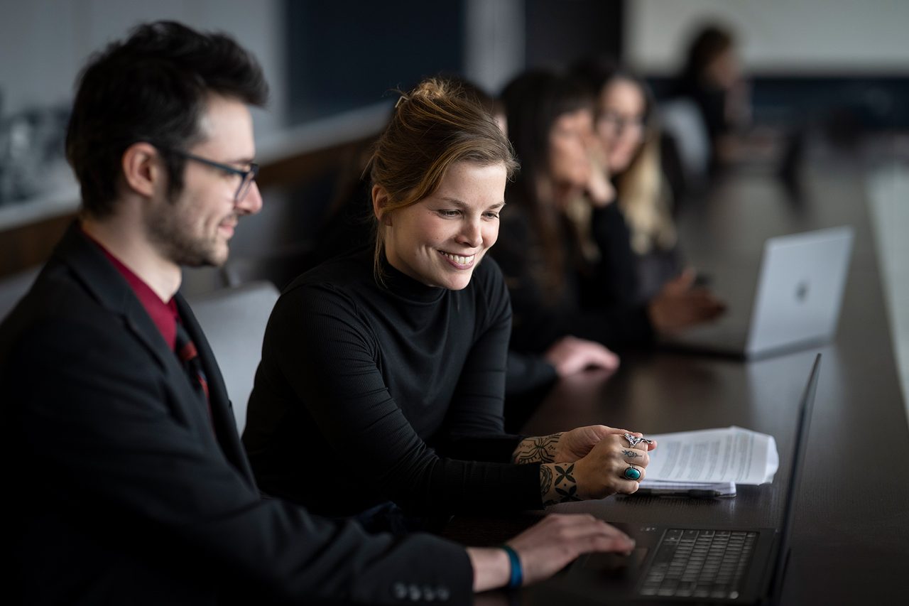 A group of professionals sit at a table with laptops, papers, and are smiling.