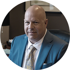 Head and shoulders portrait of a man in a blue suit with a patterned yellow tie. He's seated at a desk in an office setting.