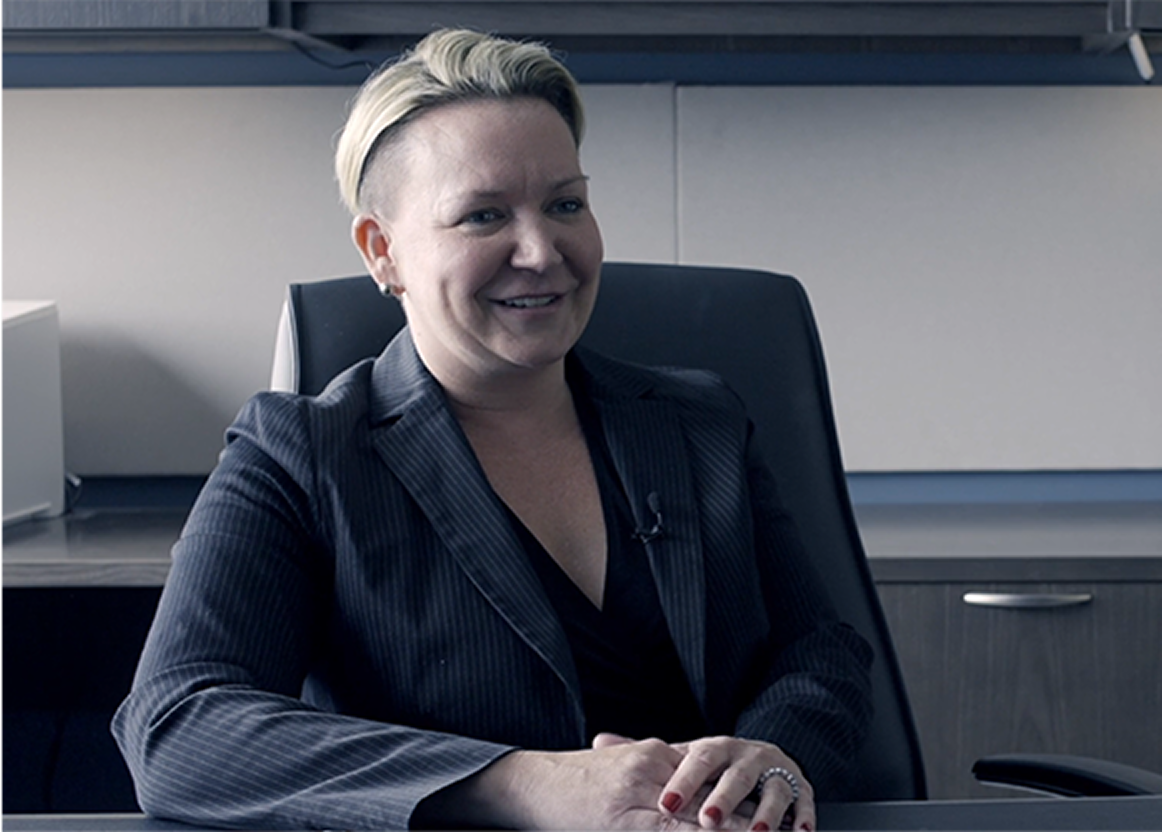 A woman in a suit smiles, seated in an office chair with her hands clasped.