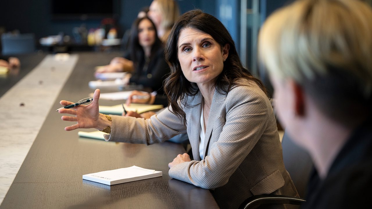 Woman in blazer gestures with pen at long conference table. Other people and notebooks are present.