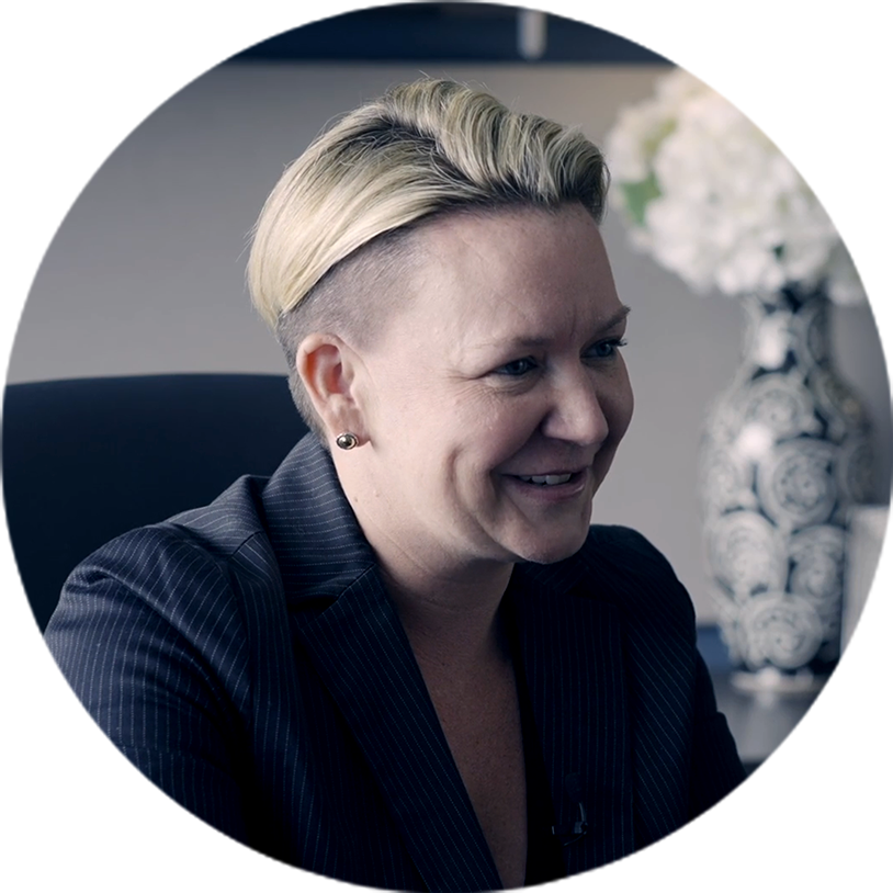 A woman with blonde hair and a dark pinstripe suit smiles slightly in an office setting.