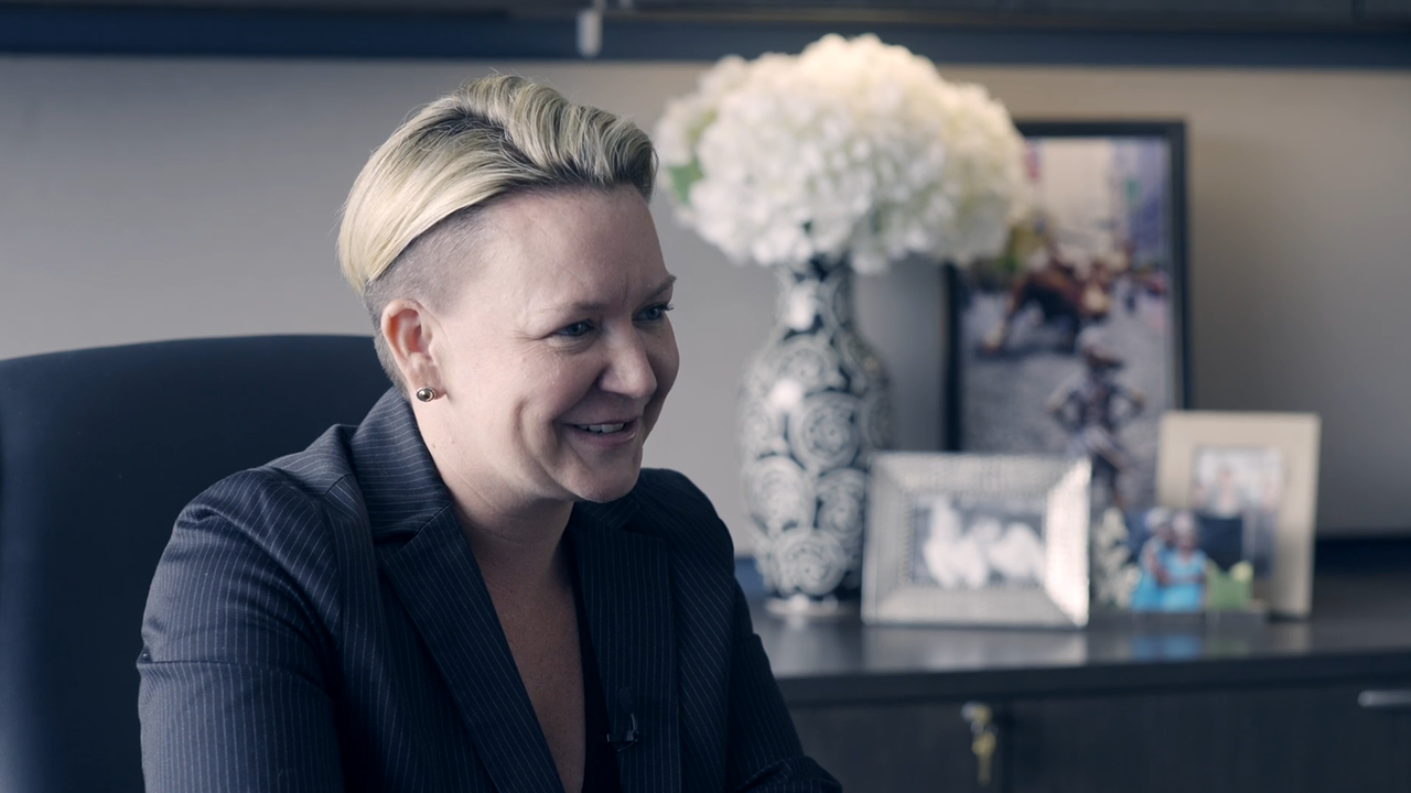 Smiling woman in pinstripe suit sits in office. White flowers and framed photos are on the desk behind.