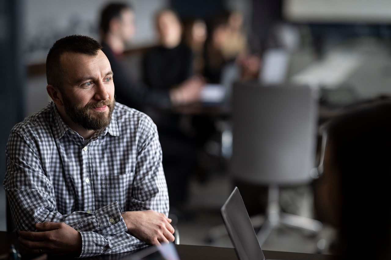 Bearded man in a checkered shirt at a meeting with others in the background.