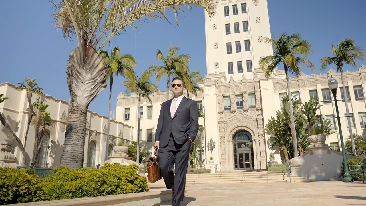 Businessman in suit walking with briefcase in front of a large, ornate building with palm trees. Sunny day.