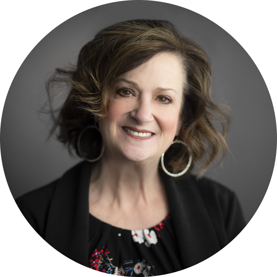 Woman with shoulder-length wavy brown hair smiles, wearing a floral blouse, black jacket, and hoop earrings against a gray...
