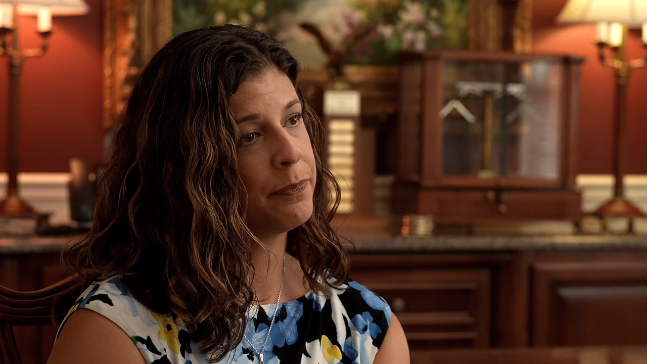 Woman with wavy brown hair, wearing a floral dress, indoors. Lamp and furniture visible in background.
