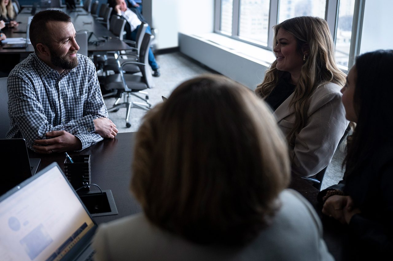 Group meeting: Four people sit at a dark table, discussing work near a window. Laptop present.