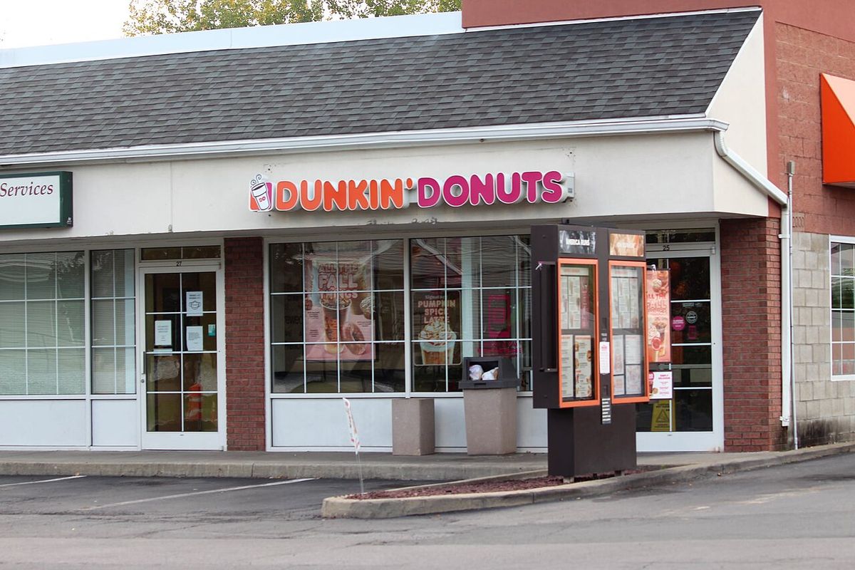A Dunkin' Donuts store in a strip mall with fall drink posters in the windows and a drive-thru menu board at the curb