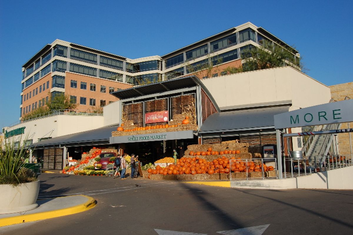 Entrance of a Whole Foods Market store decorated with hay bales and pumpkins, below a multi-story office building with blue glass windows, in Austin, Texas