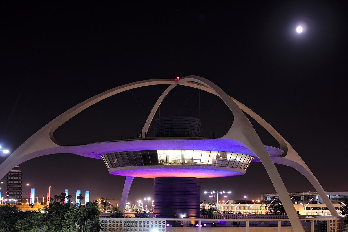 The curved white arches of the Theme Building at Los Angeles International Airport lit purple at night, with the moon in a dark sky