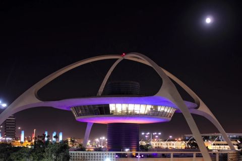The curved white arches of the Theme Building at Los Angeles International Airport lit purple at night, with the moon in a dark sky