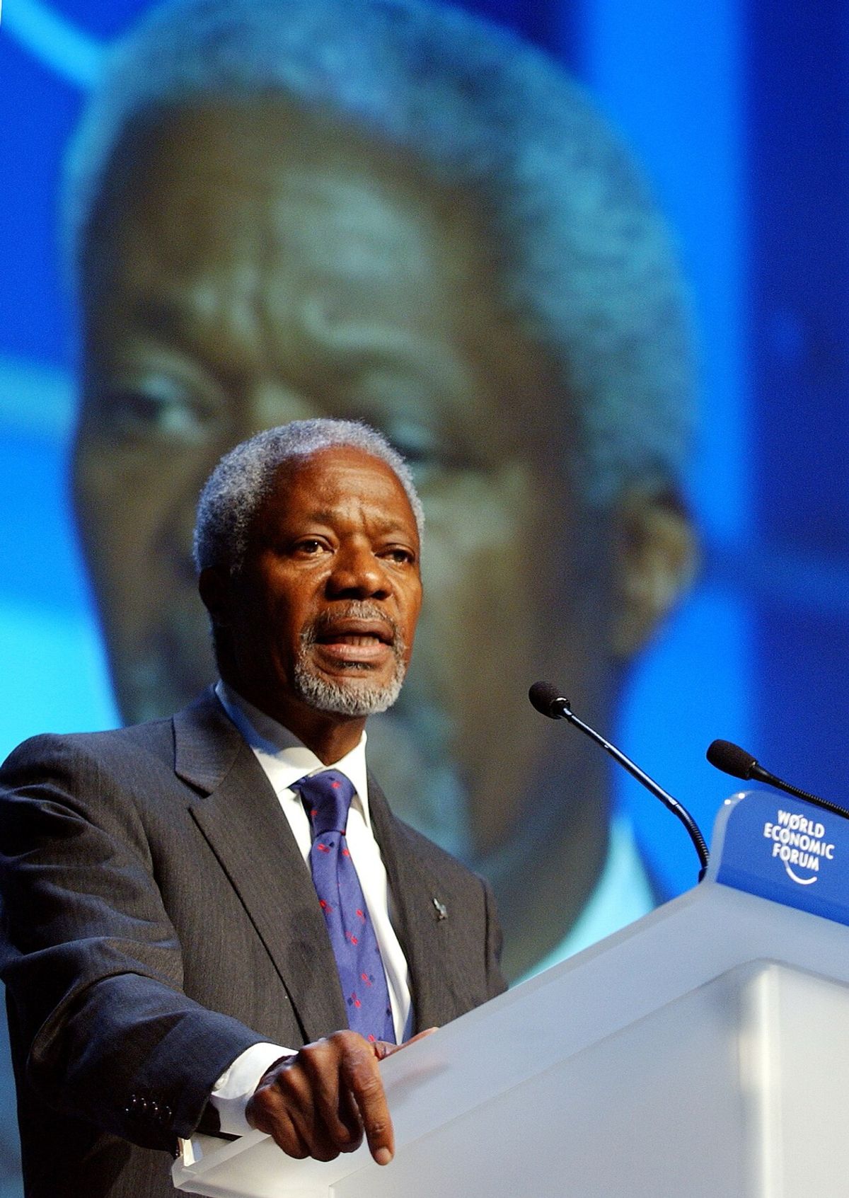 Kofi Annan speaking at a World Economic Forum lectern, with a large projection of his face on the screen behind him